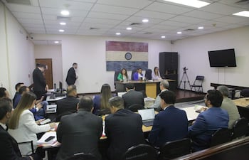 Tres mujeres en blusas formales, con bandera de Paraguay de fondo y un hombre saliendo al fondo en la reunión.