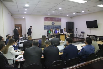 Tres mujeres en blusas formales, con bandera de Paraguay de fondo y un hombre saliendo al fondo en la reunión.