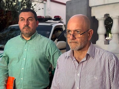 Silvio Alegre, con camisa verde, y Pablo Lemir, calvo con gafas y camisa de rayas, conversan seriamente junto a un vehículo de seguridad.o