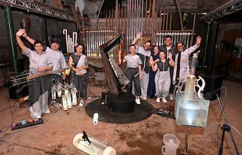 Grupo de ocho personas sonriendo en vestimenta gris y negro, junto a un gran instrumento musical en un ambiente festivo.