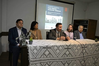 Laura Ramos con abrigo claro escucha atentamente a un hombre en traje que habla en la mesa del foro urbano, rodeado de otros participantes.