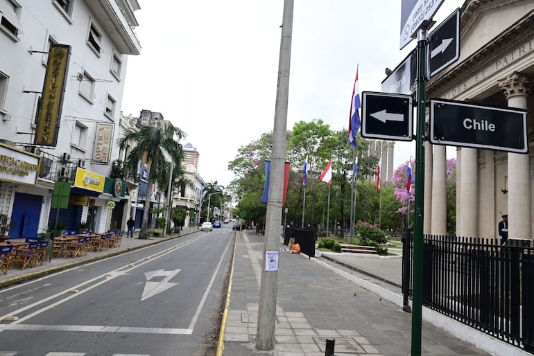 Calle desierta con locales comerciales y mesas al aire libre, rodeada de vegetación y edificios, bajo un cielo nublado.