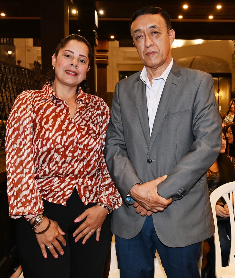 Mujer con camisa colorida y hombre con traje gris posan sonrientes en ambiente elegante rodeados de otros asistentes.