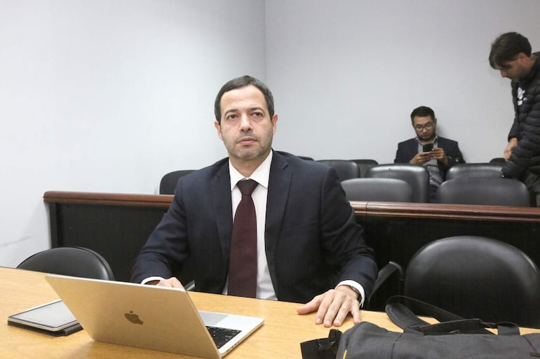 Francisco Cabrera en traje oscuro y corbata delgada, concentrado en su portátil, en un ambiente sobrio con otra persona al fondo.