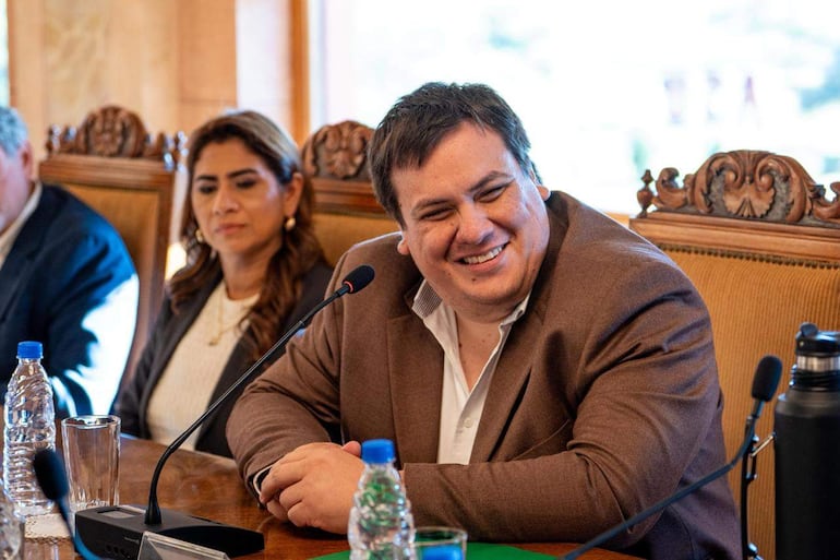 César Luis Sosa Faría sonríe mientras habla, Liz Natalia Meza Paez lo escucha atentamente. Entorno elegante con mesa y botellas de agua.