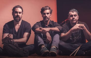 Tres hombres sentados en el suelo en un estudio, todos con camisetas oscuras y expresiones relajadas, rodeados de instrumentos musicales.