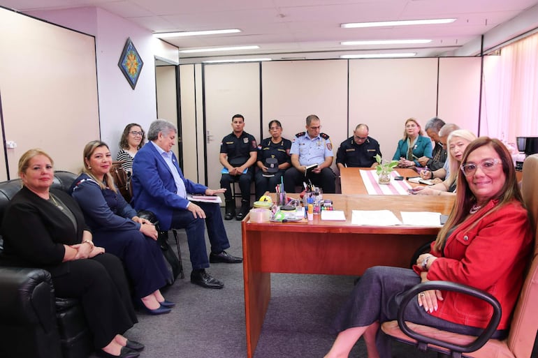 Tres mujeres sentadas en sofá negro y un hombre en traje azul discuten mientras policías uniformados observan detrás.