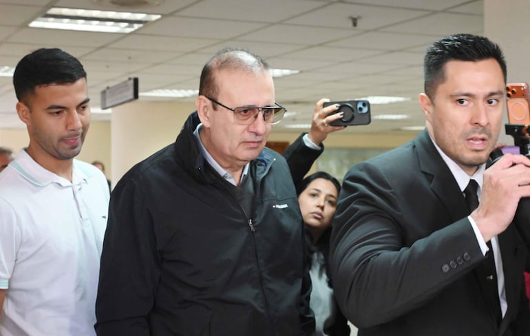 Erico Galeano Segovia con gafas y chaqueta negra, joven en camiseta blanca muestra preocupación, periodista con micrófono avanza decidido.