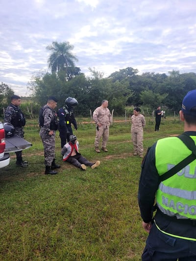 Hombre con manos atadas y chaleco rojo sentado en el suelo, rodeado de cuatro agentes de seguridad en camuflaje y chalecos negros.