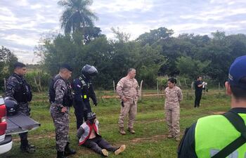 Hombre con manos atadas y chaleco rojo sentado en el suelo, rodeado de cuatro agentes de seguridad en camuflaje y chalecos negros.