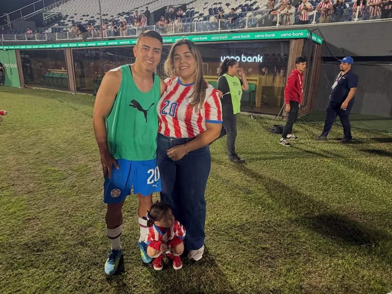 Hombre con camiseta verde y pantalones azules, sonriente, junto a un niño y una mujer con colores del equipo, en un estadio.