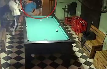 Hombre con camiseta azul siendo agredido por otro con casco en salón de villar, con gente observando en el fondo.