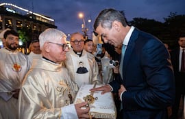 Sacerdote canoso en vestimenta dorada, Santiago Peña en traje oscuro, durante ceremonia religiosa al atardecer.