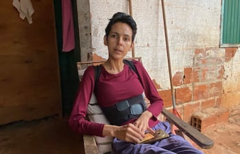 Persona de complexión delgada, con camiseta burdeos y pantalón oscuro, sentada en silla en casa, muestra postura relajada.