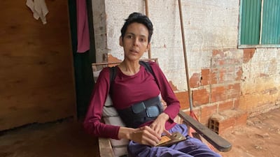 Persona de complexión delgada, con camiseta burdeos y pantalón oscuro, sentada en silla en casa, muestra postura relajada.