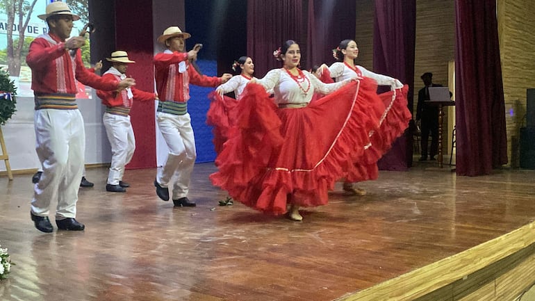 Grupo en trajes típicos rojos y blancos, bailando alegremente en un auditorio decorado.