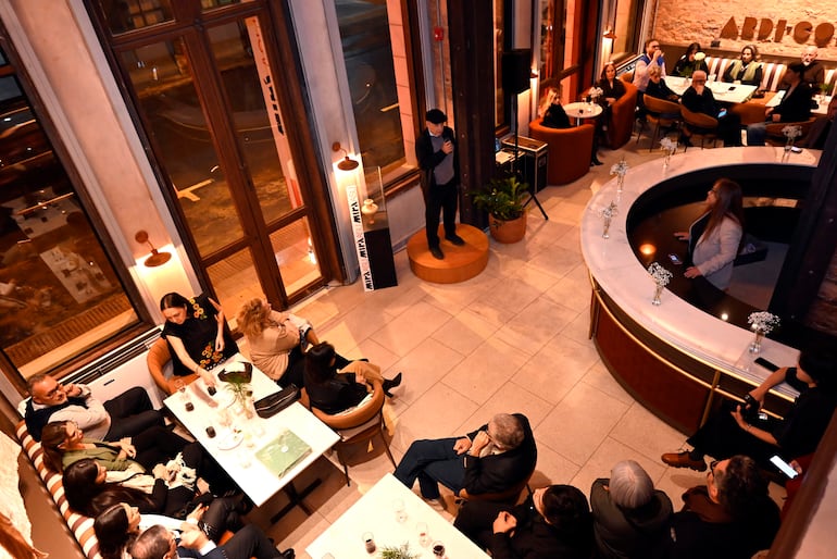 Orador masculino de pie en plataforma, público sentado en mesas con bebidas y conversando en ambiente elegante.