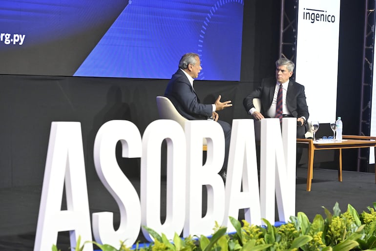 Hernando Lesme con cabello gris y traje oscuro, sorprendido, dialogando en un escenario con otra persona en traje oscuro.