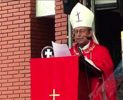 Religioso con mitra y capa roja, de pie frente a un atril en entorno de ladrillos durante ceremonia solemne.