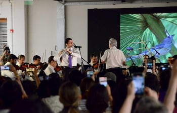 Músico joven con camisa blanca y corbata morada toca la flauta, mientras la orquesta toca bajo la dirección de un director.
