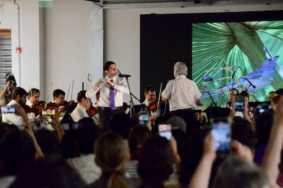 Músico joven con camisa blanca y corbata morada toca la flauta, mientras la orquesta toca bajo la dirección de un director.