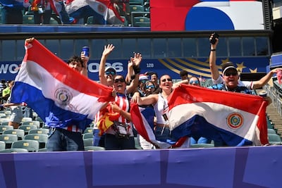 Seis aficionados sostienen banderas de Paraguay enérgicamente, con expresiones felices en un estadio lleno de asientos y una pantalla al fondo.