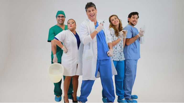 Grupo de cinco personas con atuendos médicos y sonrisas, en un estudio con fondo blanco, sosteniendo jeringa y utensilio médico.