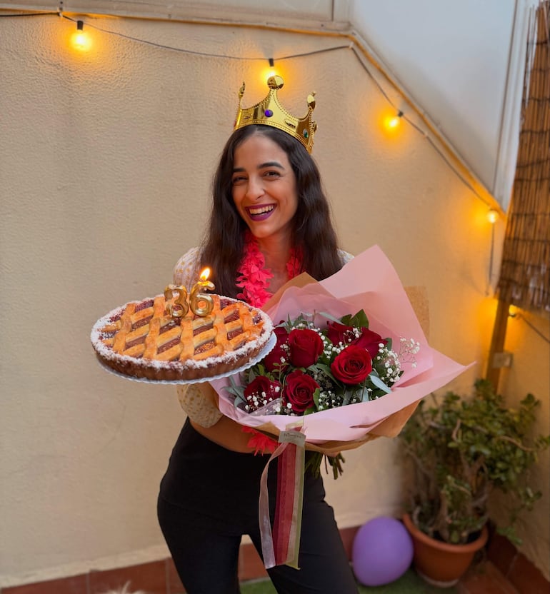 Mujer sonriente con corona dorada, sosteniendo una tarta con vela y ramo de rosas en un entorno iluminado.