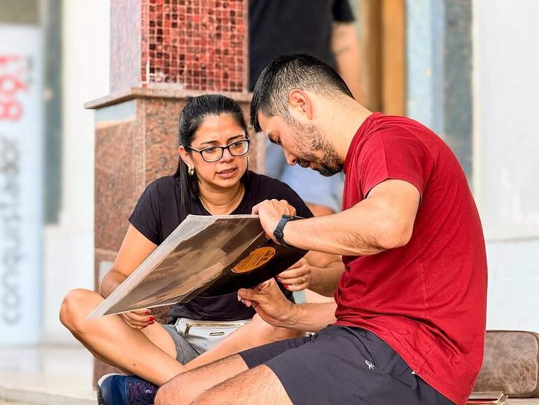 Mujer con camiseta negra y anteojos conversa con hombre en camiseta roja, ambos revisan un disco en ambiente al aire libre.