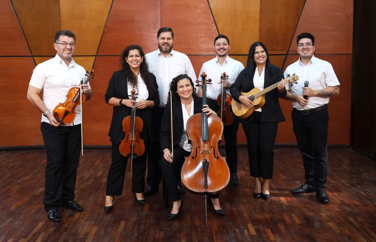 Siete músicos sonrientes en camisas blancas y pantalones oscuros, sosteniendo instrumentos como violonchelo y violín en un fondo cálido.