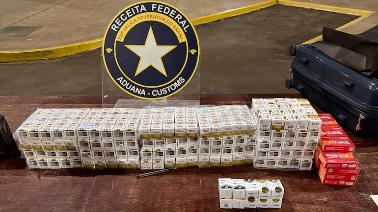 Cartel de Receita Federal con cigarrillos confiscados en una mesa, maleta negra al lado, sin personas visibles.