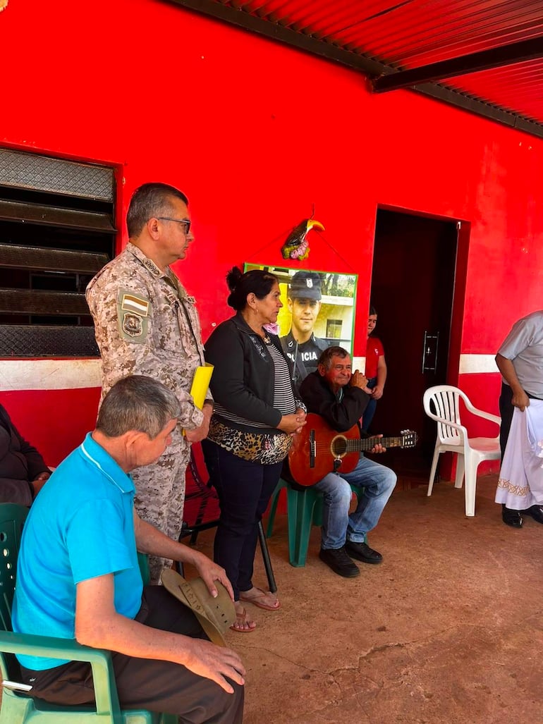 Mujer en blusa negra habla frente a público, rodeada de hombres, con un retrato de policía de fondo y un ambiente emotivo.