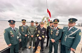 Siete militares en uniforme, uno en blanco destaca, mientras Maruca, la cachorra, observa con tranquilidad en un bote sobre el agua.