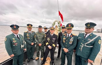 Siete militares en uniforme, uno en blanco destaca, mientras Maruca, la cachorra, observa con tranquilidad en un bote sobre el agua.