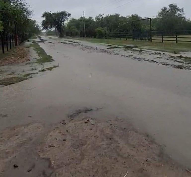 Paisaje inundado con agua cubriendo la calle, cercas y vegetación visible, bajo un cielo nublado.