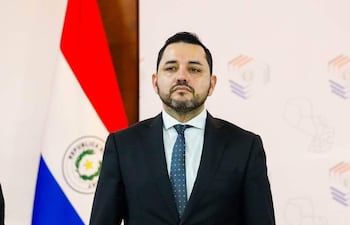 Luis Bello con traje oscuro, Jorge Bogarín González a su lado, ambos en conferencia rodeados de banderas paraguayas y documentos.