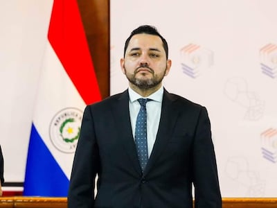 Luis Bello con traje oscuro, Jorge Bogarín González a su lado, ambos en conferencia rodeados de banderas paraguayas y documentos.