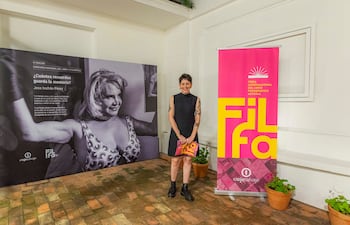 Mujer de pie en vestido negro sosteniendo un libro colorido, con cartel de FILFA y plantas de decoración en el fondo.