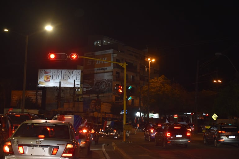 Escena nocturna con vehículos detenidos en un semáforo rojo, luces brillantes y banderas de Paraguay visibles en el ambiente urbano.