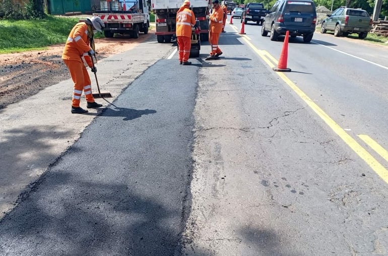 Tres trabajadores con overoles naranjas y cascos asfaldan la carretera, mientras un camión y autos están estacionados al fondo.