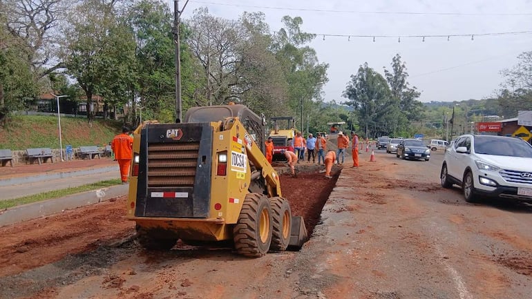 Grupo de trabajadores en ropa naranja supervisa trabajos de construcción en la carretera, con un vehículo CAT en el lugar.
