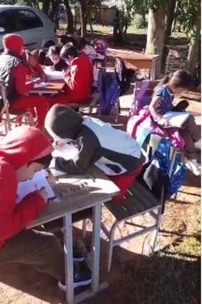 Niños con abrigo y gorros escriben en cuadernos en mesas al aire libre, rodeados de árboles.