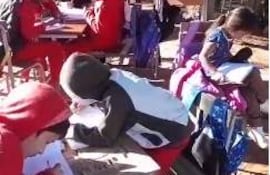 Niños con abrigo y gorros escriben en cuadernos en mesas al aire libre, rodeados de árboles.