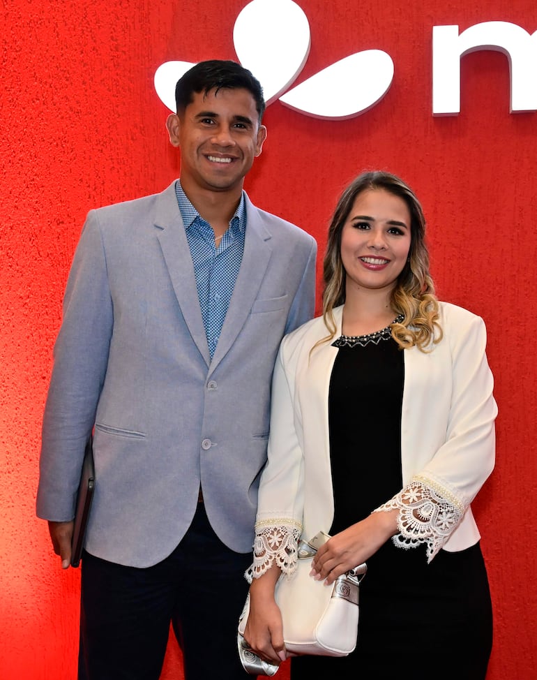 Hombre en blazer claro y mujer en blusa negra y abrigo blanco sonríen ante fondo rojo con logotipo institucional.