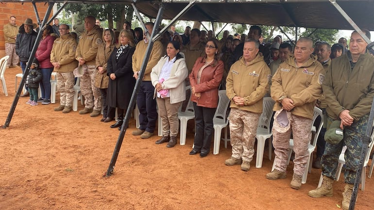Grupo de personas en torno a una celebración, vistiendo tonos beige y camuflaje, en un ambiente solemne al aire libre.
