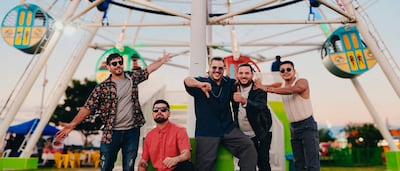Cinco hombres sonrientes en parque de diversiones, vestidos con ropa colorida, disfrutando del ambiente festivo y la rueda de la fortuna al fondo.