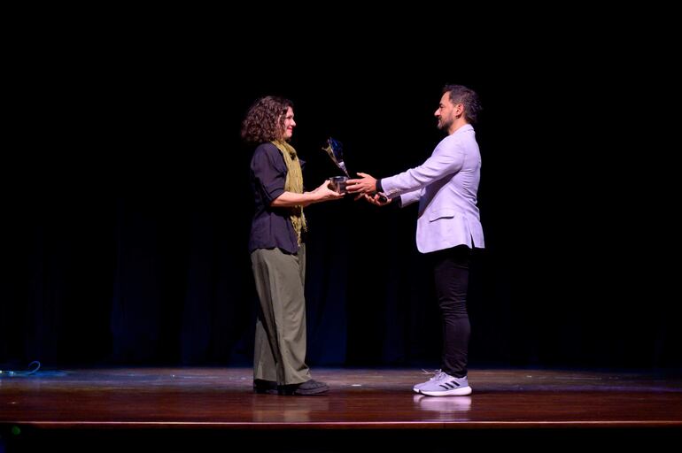 Mujer con cabello rizado y blusa negra recibe un trofeo de hombre en blazer blanco, fondo oscuro y luz suave.