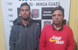 Dos hombres, uno con chaqueta oscura y otro con suéter rojo, lucen serios frente a la comisaría de Minga Guazú.
