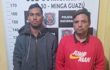 Dos hombres, uno con chaqueta oscura y otro con suéter rojo, lucen serios frente a la comisaría de Minga Guazú.