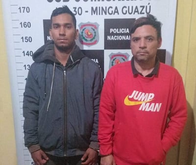 Dos hombres, uno con chaqueta oscura y otro con suéter rojo, lucen serios frente a la comisaría de Minga Guazú.
